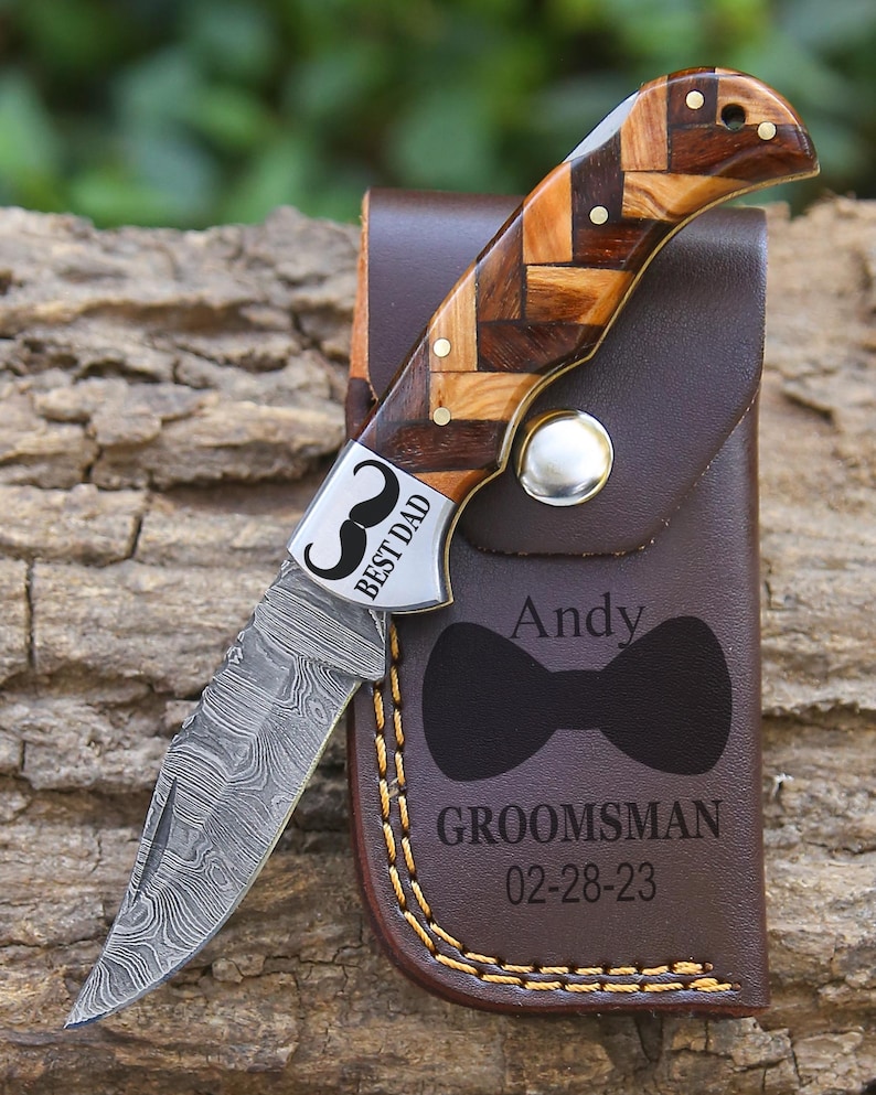 May include: A folding knife with a Damascus steel blade and a handle featuring a brown wood herringbone pattern. The bolster is engraved with a mustache and the text BEST DAD. It is paired with a brown leather sheath engraved with a bow tie, the name Andy, the word GROOMSMAN, and the date 02-28-23.