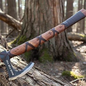 May include: A handcrafted axe with a wooden handle and a dark metal blade. The handle is wrapped with leather and features etched symbols. The blade has the text "MADE FOR YOU". The axe is resting on a log in a forest setting.