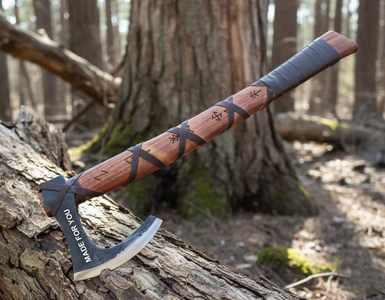 May include: A handcrafted axe with a wooden handle and a dark metal blade. The handle is wrapped with leather and features etched symbols. The blade has the text "MADE FOR YOU". The axe is resting on a log in a forest setting.