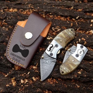 May include: A brown leather knife case with a silver snap closure, a black bow tie graphic, and the text "Andy GROOMSMAN 02-28-23". Two folding knives with patterned blades and horn handles are also visible. One knife has "BEST DAD" text.