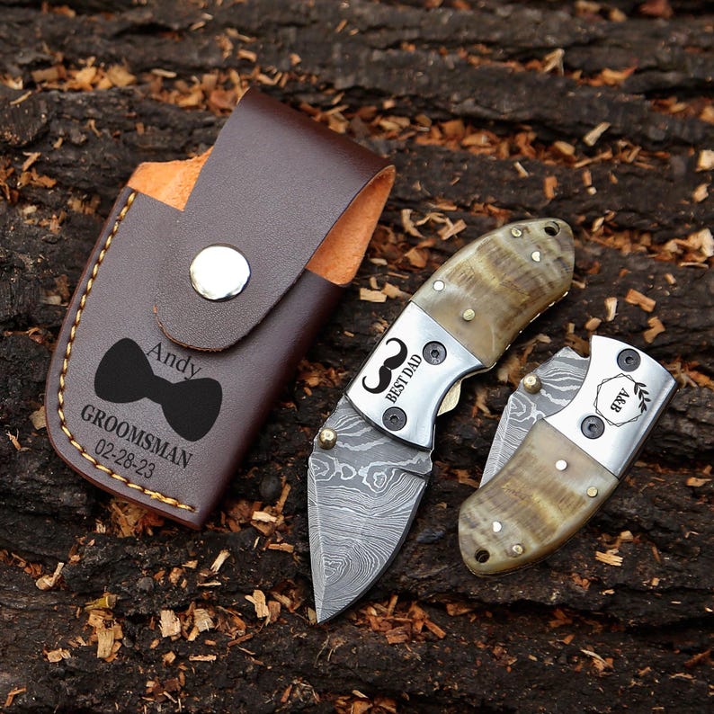 May include: A brown leather knife case with a silver snap closure, a black bow tie graphic, and the text "Andy GROOMSMAN 02-28-23". Two folding knives with patterned blades and horn handles are also visible. One knife has "BEST DAD" text.