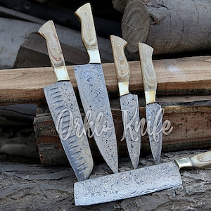 May include: A set of five kitchen knives with light wood handles and patterned steel blades. The blades have a unique design, and the handles are secured with brass bolsters. The knives are arranged on a wooden surface, with the text "Orklo Knife" visible.