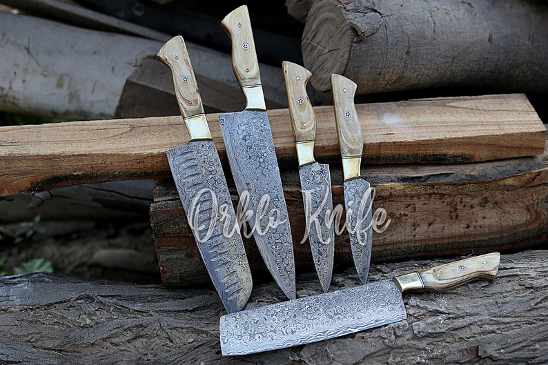May include: A set of five kitchen knives with light wood handles and patterned steel blades. The blades have a unique design, and the handles are secured with brass bolsters. The knives are arranged on a wooden surface, with the text "Orklo Knife" visible.