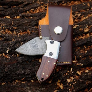 May include: A small, folding pocket knife with a patterned steel blade and a dark wood handle. The knife is partially open, revealing the blade's intricate design. It rests next to a brown leather sheath with a snap closure.