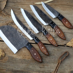 May include: An assortment of handcrafted knives with dark, textured blades and wooden handles. The blades have a unique, rustic appearance, and the handles are a rich brown color. The knives are arranged on a weathered wooden surface.
