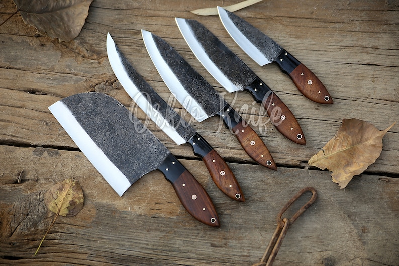 May include: An assortment of handcrafted knives with dark, textured blades and wooden handles. The blades have a unique, rustic appearance, and the handles are a rich brown color. The knives are arranged on a weathered wooden surface.
