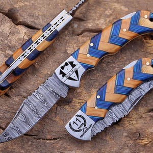May include: Three folding knives with Damascus steel blades and handles featuring a blue and tan herringbone pattern. One knife is open, while two are closed. The open knife features a tuxedo graphic with the text Best Man, and another knife displays a laurel wreath graphic with the text 11 Years Anniversary.