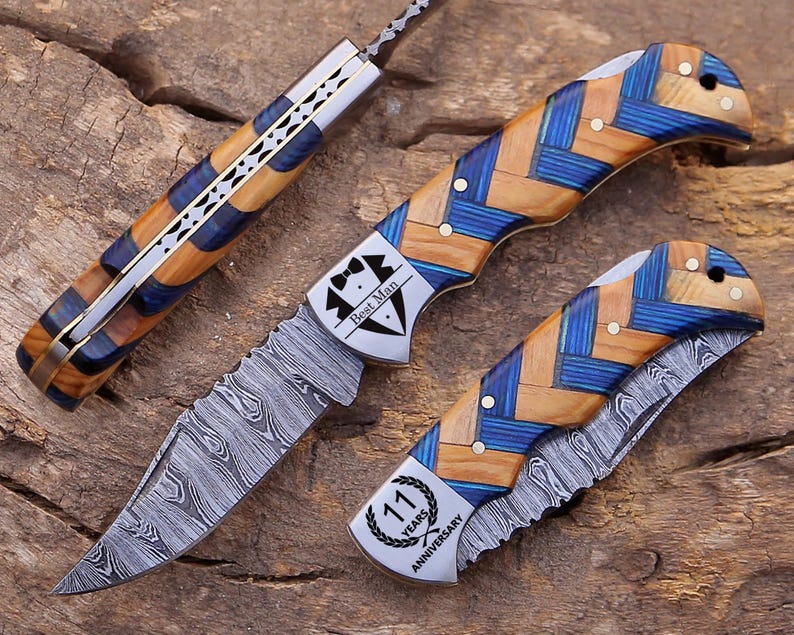 May include: Three folding knives with Damascus steel blades and handles featuring a blue and tan herringbone pattern. One knife is open, while two are closed. The open knife features a tuxedo graphic with the text Best Man, and another knife displays a laurel wreath graphic with the text 11 Years Anniversary.