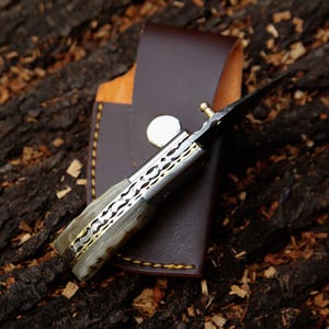 May include: A handcrafted folding knife with a decorative handle and a sharp blade, partially inserted into a brown leather sheath. The knife features a patterned metal design and a brass accent. The sheath has a snap closure and stitched edges.