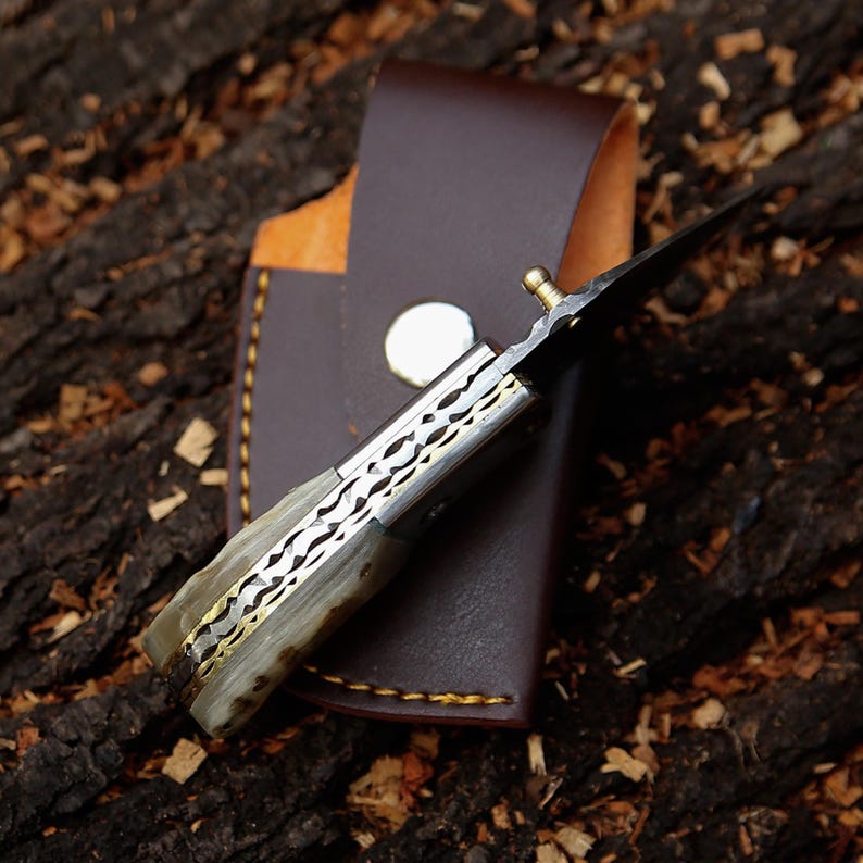 May include: A handcrafted folding knife with a decorative handle and a sharp blade, partially inserted into a brown leather sheath. The knife features a patterned metal design and a brass accent. The sheath has a snap closure and stitched edges.