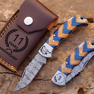 May include: Two folding knives with Damascus steel blades and handles featuring a blue and tan woven pattern. One knife is open, the other closed. A brown leather sheath is engraved with a laurel wreath and the text 11 YEARS ANNIVERSARY.