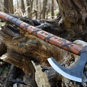 May include: A handcrafted axe with a wooden handle and a curved, silver-coloured blade. The handle is wrapped with dark leather straps and features etched runes. The axe is resting on a weathered wooden surface, suggesting a rustic or historical theme.