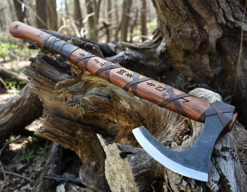 May include: A handcrafted axe with a wooden handle and a curved, silver-coloured blade. The handle is wrapped with dark leather straps and features etched runes. The axe is resting on a weathered wooden surface, suggesting a rustic or historical theme.