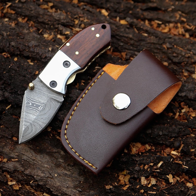 May include: A small folding knife with a patterned blade and a wooden handle, next to a brown leather sheath. The knife has a silver bolster and brass rivets. The sheath has a snap closure and yellow stitching.
