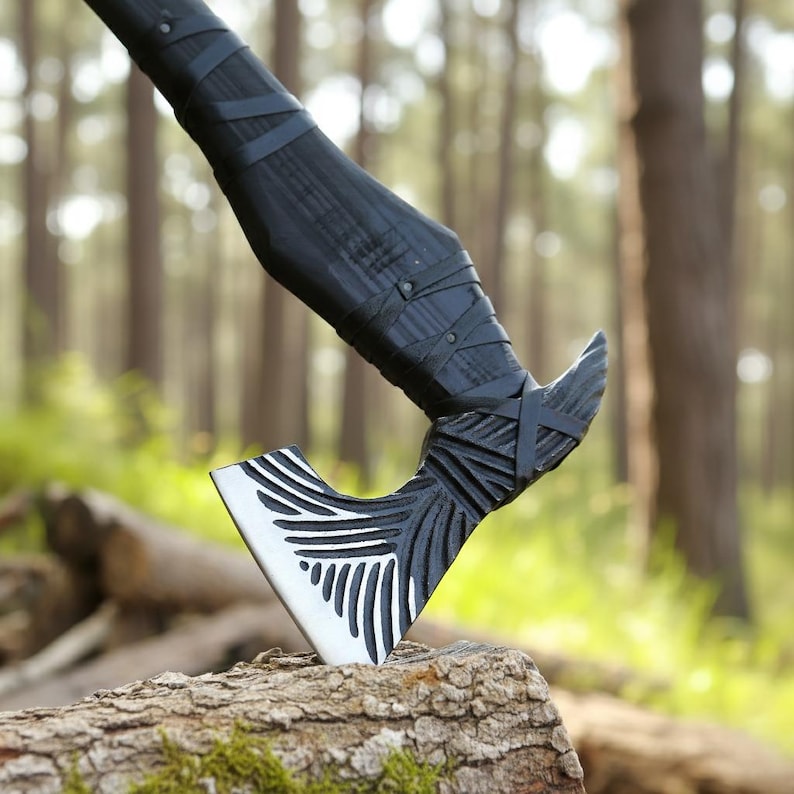 May include: A black and silver axe with a detailed blade design and a wrapped handle. The axe head features a white blade with black linear patterns. The handle is wrapped in black leather. The axe is resting on a piece of wood.