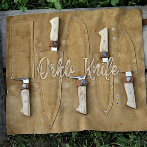 May include: A set of knives with light wood handles and leather sheaths, displayed on a tan leather roll. The knives are secured with brown leather straps. The text "Orklo Knife" is visible.