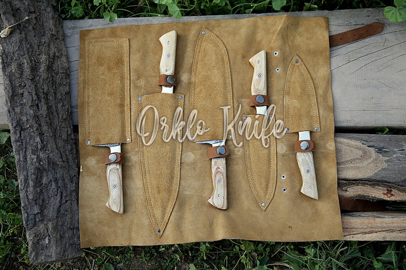 May include: A set of knives with light wood handles and leather sheaths, displayed on a tan leather roll. The knives are secured with brown leather straps. The text "Orklo Knife" is visible.