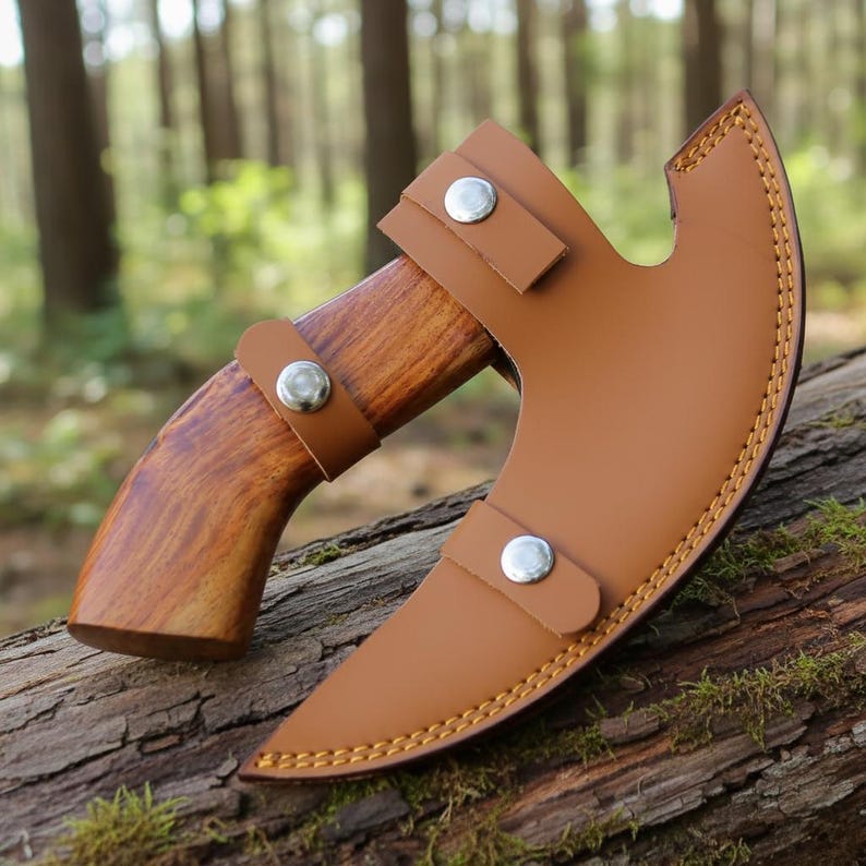 May include: A small axe with a wooden handle and a brown leather sheath. The axe head is curved and the sheath is secured with two leather straps and silver-coloured metal studs. The axe is resting on a log in a forest setting.