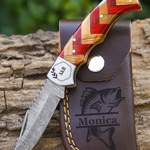 May include: A folding pocket knife with a Damascus steel blade and a handle featuring a red and tan woven wood pattern. The metal bolster is engraved with the text A&B and a leaf icon. It is paired with a brown leather sheath featuring a fish graphic and the name Monica.