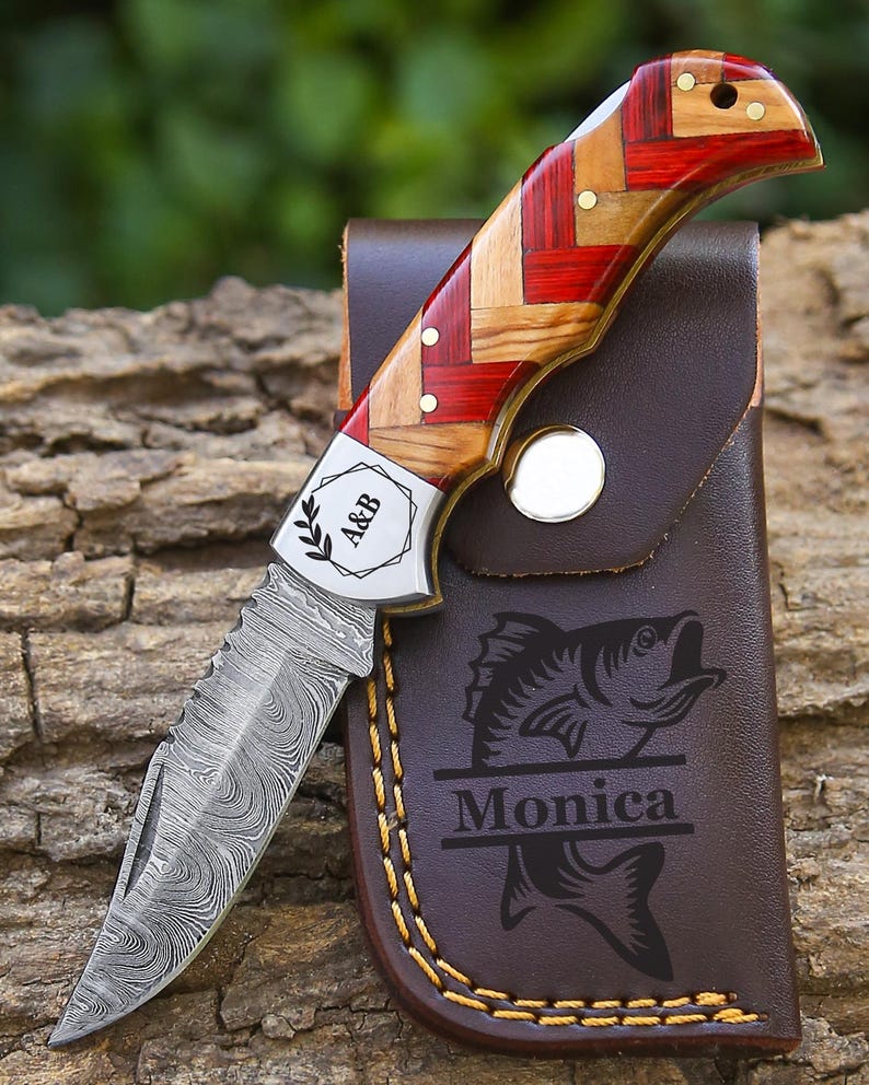 May include: A folding pocket knife with a Damascus steel blade and a handle featuring a red and tan woven wood pattern. The metal bolster is engraved with the text A&B and a leaf icon. It is paired with a brown leather sheath featuring a fish graphic and the name Monica.