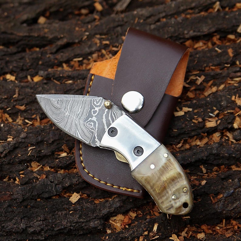 May include: A small, folding knife with a patterned blade and a horn handle. The knife is paired with a brown leather sheath. The blade has a unique, swirling pattern. The handle is a light brown color. The sheath is a darker brown.