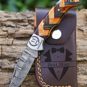 May include: A folding pocket knife with a Damascus steel blade and a multi-colored wood handle featuring a metal bolster engraved with 11 Years Anniversary. It is paired with a brown leather sheath embossed with a tuxedo and Best Man text.