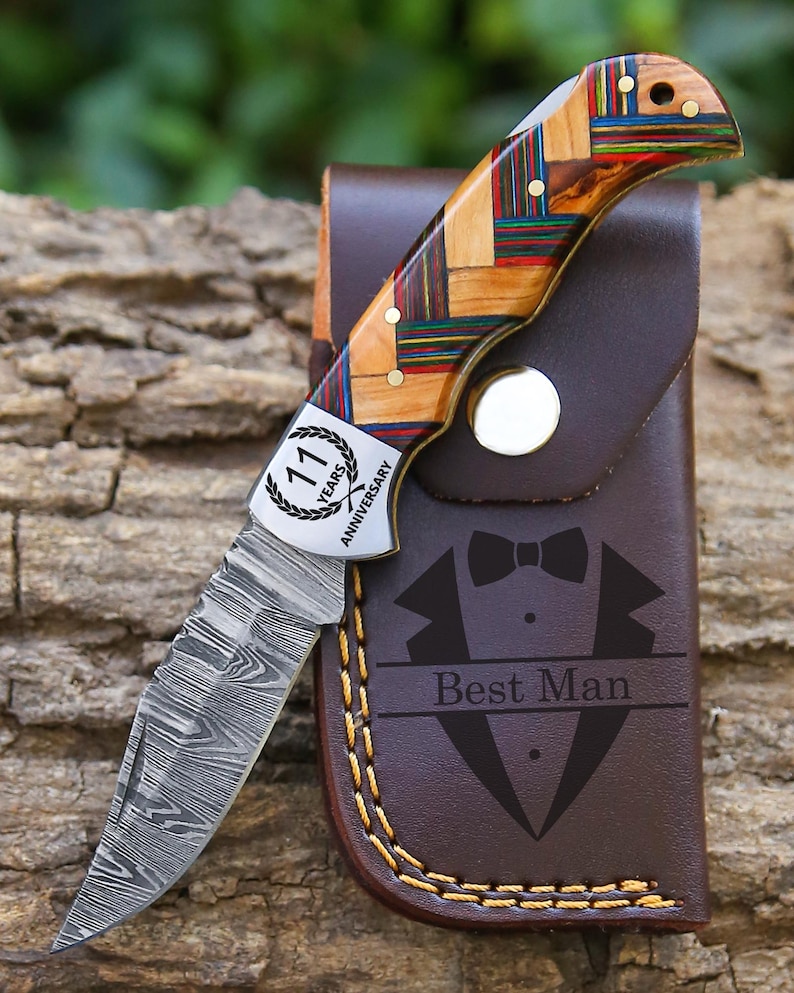May include: A folding pocket knife with a Damascus steel blade and a multi-colored wood handle featuring a metal bolster engraved with 11 Years Anniversary. It is paired with a brown leather sheath embossed with a tuxedo and Best Man text.