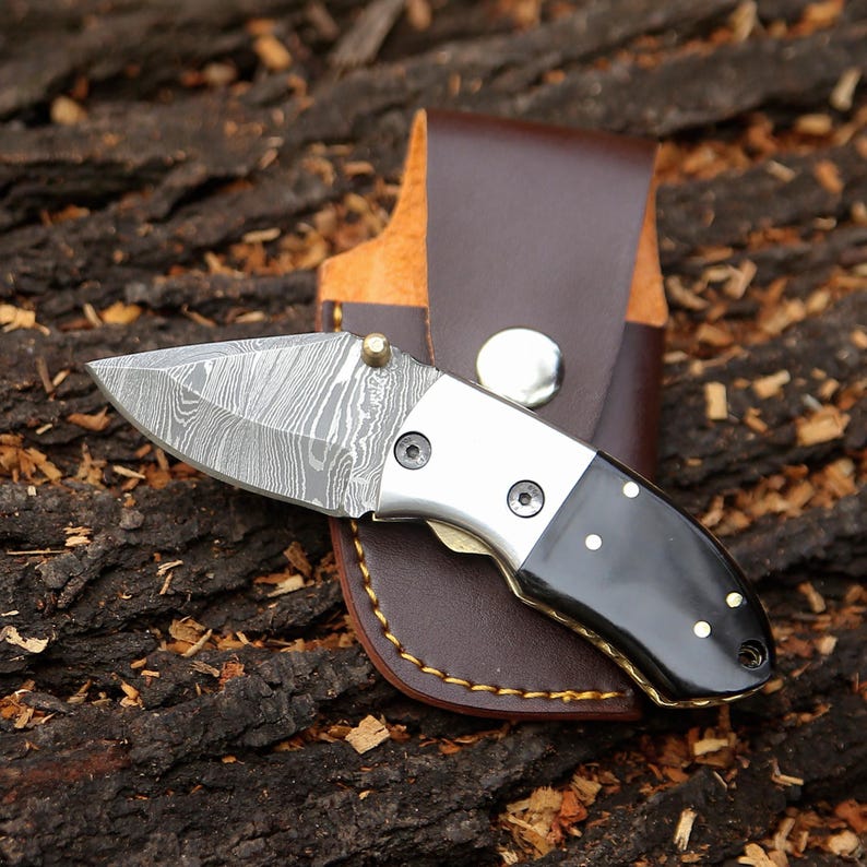May include: A small folding knife with a patterned blade and a black and silver handle. The knife is in a brown leather sheath. The blade has a unique pattern. The handle has brass accents. The knife is designed for everyday carry.