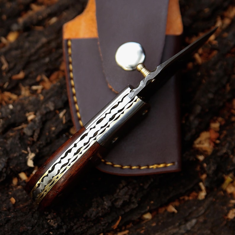May include: A handcrafted knife with a dark wooden handle, silver and gold accents, and a sharp, black blade. The knife is partially sheathed in a brown leather case with a silver button closure. The knife is designed for utility and collection.
