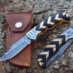 May include: Two folding knives with Damascus steel blades and herringbone patterned wood handles. One knife is open, showing the text "LY Love You" on the bolster. The other is closed, showing "Your Date / Text" on the bolster. A brown leather sheath is included.