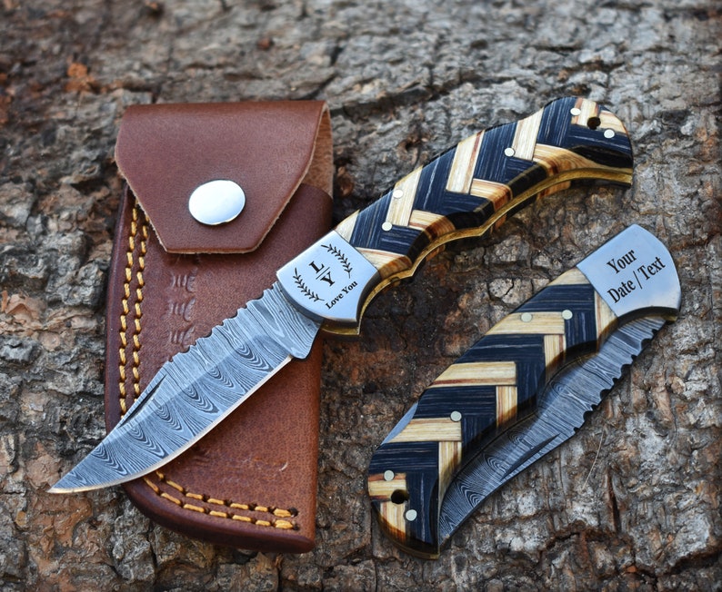 May include: Two folding knives with Damascus steel blades and herringbone patterned wood handles. One knife is open, showing the text "LY Love You" on the bolster. The other is closed, showing "Your Date / Text" on the bolster. A brown leather sheath is included.