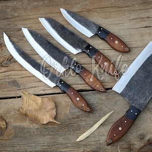 May include: A collection of handcrafted knives with dark, textured blades and wooden handles. The blades have a light-colored edge, and the handles are a rich brown with black accents. The knives are arranged on a rustic wooden surface.