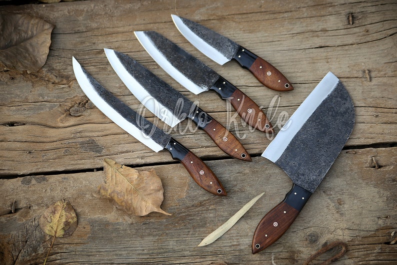 May include: A collection of handcrafted knives with dark, textured blades and wooden handles. The blades have a light-colored edge, and the handles are a rich brown with black accents. The knives are arranged on a rustic wooden surface.