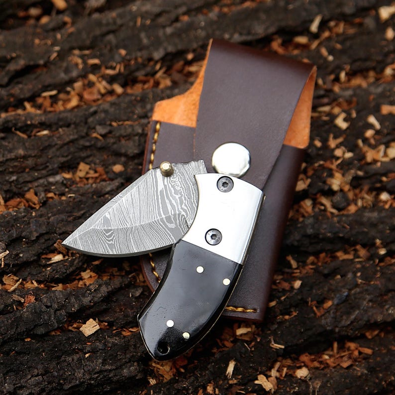 May include: A small, open folding knife with a patterned blade and a black handle with brass accents. The knife is paired with a brown leather sheath. The blade is made of steel. The knife is resting on a wood surface.