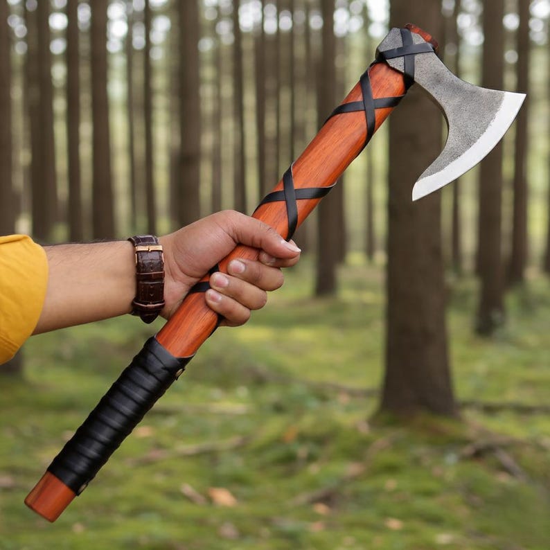 May include: A handcrafted axe with a wooden handle and a steel blade. The handle is wrapped with black leather and features a crisscross design. The axe is held against a backdrop of a forest.