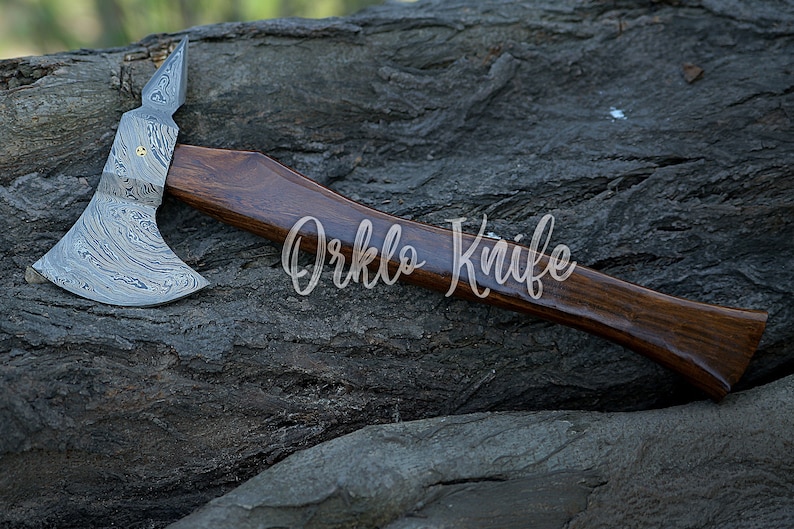 May include: A handcrafted axe with a dark wooden handle and a patterned steel blade. The axe head features a unique design with a pointed top. The text "Orklo Knife" is visible on the handle.