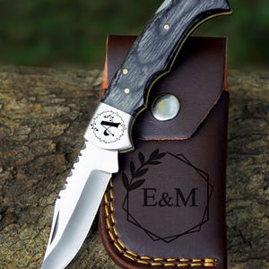 May include: A folding pocket knife with a silver blade and a black and gray wooden handle. The knife is resting on a brown leather sheath with the initials "E&M" and a decorative leaf design. The knife is on a wooden surface.