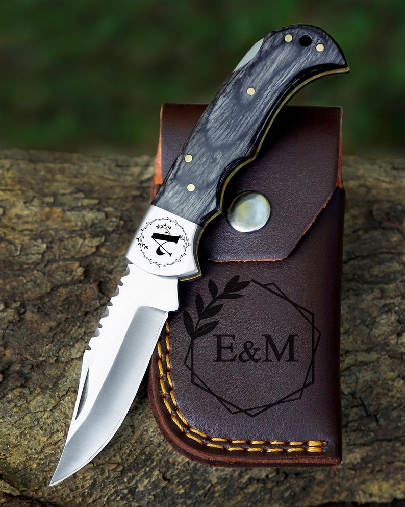 May include: A folding pocket knife with a silver blade and a black and gray wooden handle. The knife is resting on a brown leather sheath with the initials "E&M" and a decorative leaf design. The knife is on a wooden surface.