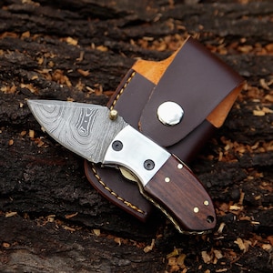 May include: A small, folding pocket knife with a patterned blade and a wooden handle. The knife has a silver bolster and brass rivets. It is shown with a brown leather sheath with a snap closure.
