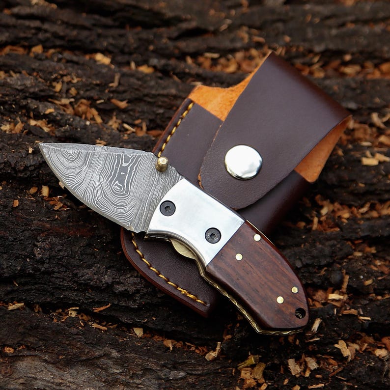 May include: A small, folding pocket knife with a patterned blade and a wooden handle. The knife has a silver bolster and brass rivets. It is shown with a brown leather sheath with a snap closure.
