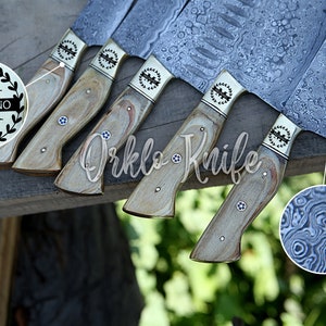 May include: A collection of knives with patterned steel blades and light wood handles. Each knife has a decorative metal bolster and a small, circular inlay. The blades have a unique, swirling pattern. The text "Orklo Knife" is visible.