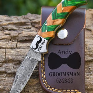 May include: A pocket knife with a patterned blade and a handle made of wood and green accents. The knife is in a brown leather sheath with the text "Andy", a bow tie graphic, and "GROOMSMAN 02-28-23". The knife has the text "BEST DAD".