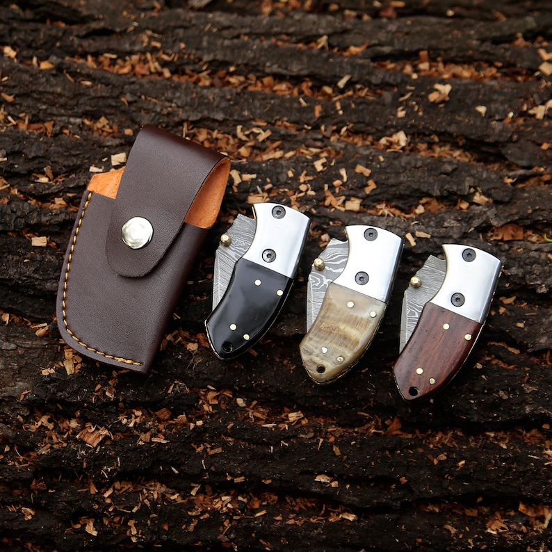 May include: A brown leather sheath with a silver snap button next to three small folding knives. The knives have silver blades and handles made of black, tan, and brown materials. The knives are on a textured brown surface.