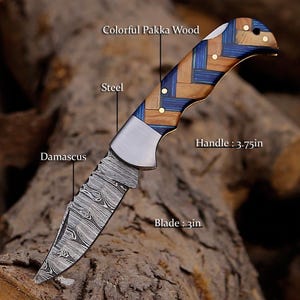 May include: A folding knife with a 3-inch Damascus steel blade and a 3.75-inch handle made of colorful Pakka wood in a blue and brown woven pattern. The knife features a steel bolster and brass pins, with text labels identifying the blade, steel, handle, and wood.