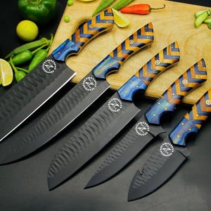 May include: Set of six black kitchen knives with patterned wooden handles in blue, yellow, and brown. The blades have a textured finish. The knives are arranged on a wooden cutting board with vegetables.