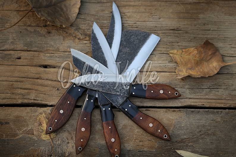 May include: A collection of handcrafted knives with dark wooden handles and textured blades arranged in a circular pattern. The blades have a silver finish, and the handles are a rich brown with decorative rivets. The knives are displayed on a rustic wooden surface.
