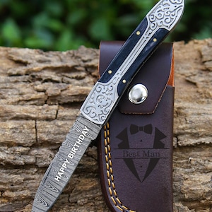 May include: A folding knife with a decorative handle and blade, engraved with "HAPPY BIRTHDAY." The knife has a black and silver handle with gold accents. It rests in a brown leather sheath with "Best Man" text and a bow tie graphic.