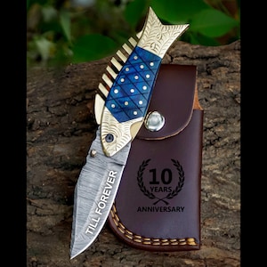 May include: A folding knife with a fish-shaped handle featuring a blue and gold design. The blade is engraved with "TILL FOREVER." A brown leather sheath with "10 YEARS ANNIVERSARY" is also included.
