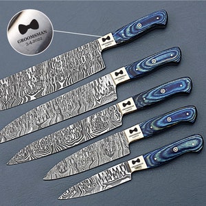 May include: A set of five Damascus steel knives with blue and black wood grain handles. Each knife has a unique blade pattern and a silver bolster. The handles are a mix of blue and black. The text "GROOMSMAN 3-4-2022" is engraved on the bolster.