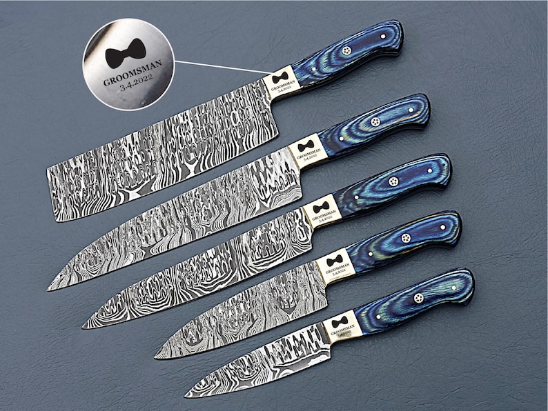 May include: A set of five Damascus steel knives with blue and black wood grain handles. Each knife has a unique blade pattern and a silver bolster. The handles are a mix of blue and black. The text "GROOMSMAN 3-4-2022" is engraved on the bolster.